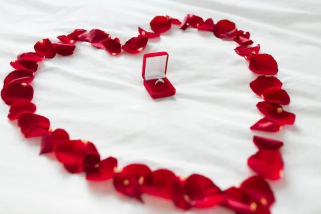 Red ring box with diamond ring inside, surrounded by heart petals in the shape of a heart on bed sheets
