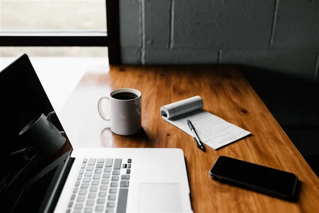 Image of a table with a laptop, coffee cut, pad of paper and phone laid beside it