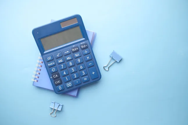 A blue calculator sits on top of a small spiral-bound notebook, surrounded by two blue binder clips, against a light blue background