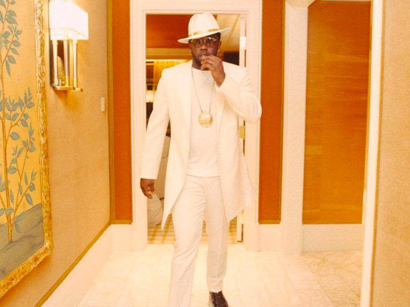 P. Diddy wearing an all-white outfit, walking through a lavish hotel suite with gold and wood accents