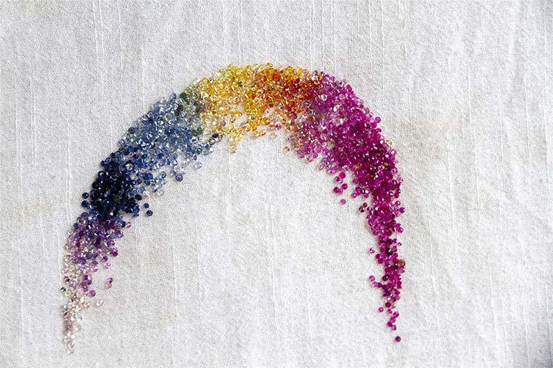 Different-colored loose sapphires, arranged like a rainbow against a textured white background