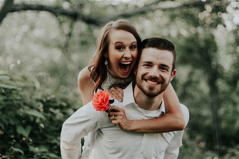 A joyful woman piggybacking a smiling man, both dressed in white, suggesting a wedding celebration