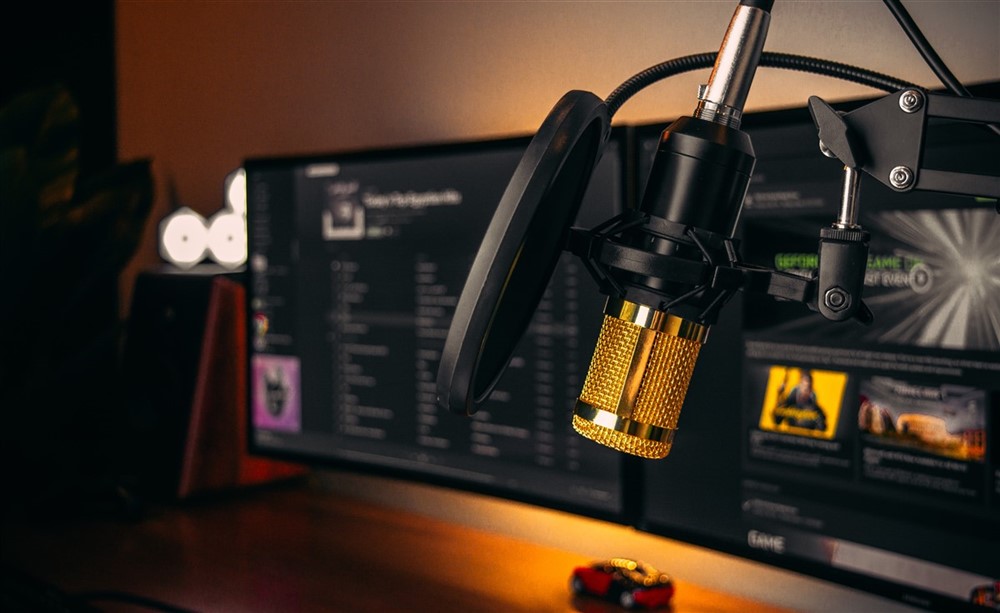 Close-up of a gold microphone in front of a dual computer screen setup with a pop filter