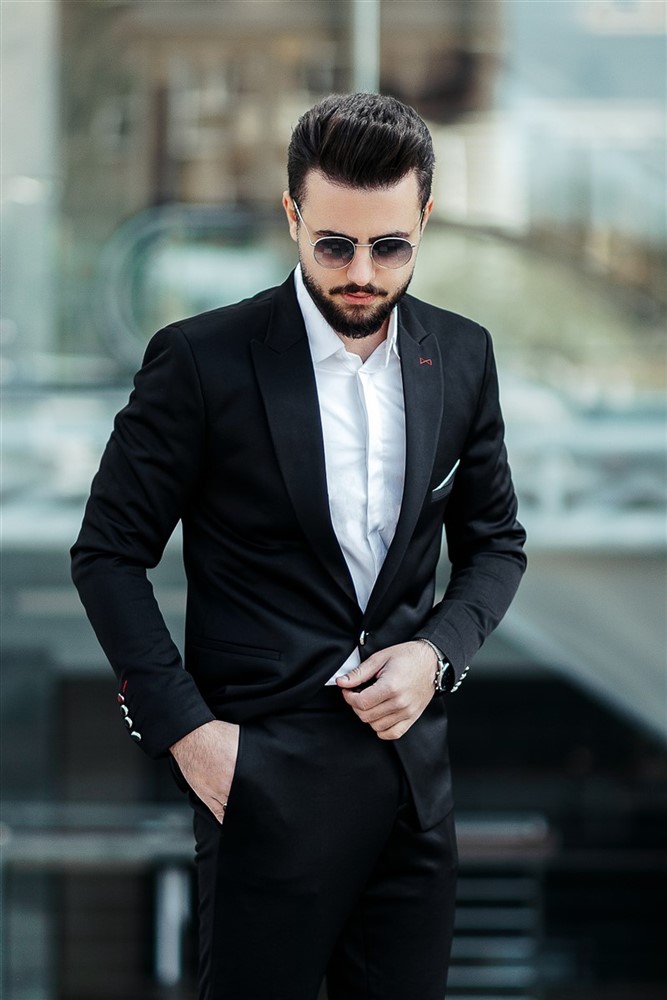 A stylish man in a sleek black suit and sunglasses stands confidently, hands in pockets, against an urban backdrop