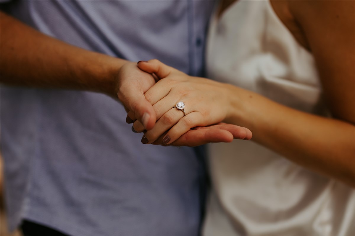 A close-up of a couple holding hands, with a focus on the woman's hand showcasing an elegant engagement ring