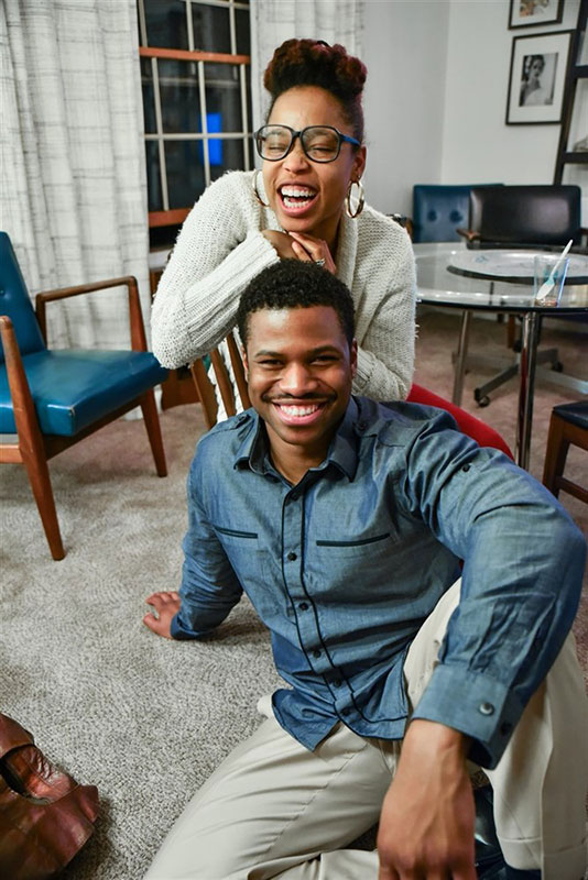 A woman sits on a chair while a man sits on the floor in front of her; both are smiling warmly, conveying happiness