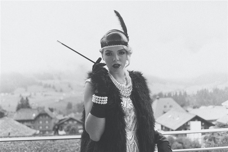 A woman dressed in 1920s flapper fashion, wearing a feathered headband, pearls, and a fur stole, holding a cigarette holder and exuding vintage elegance