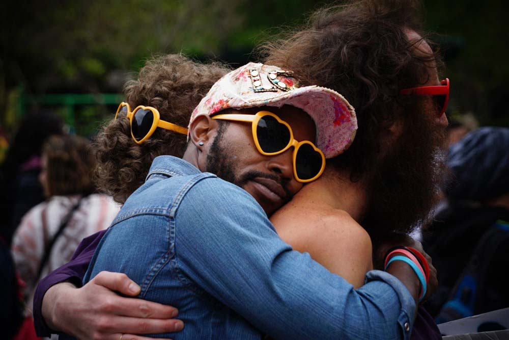 Three men hugging warmly, all wearing heart-shaped sunglasses, at an outdoor event