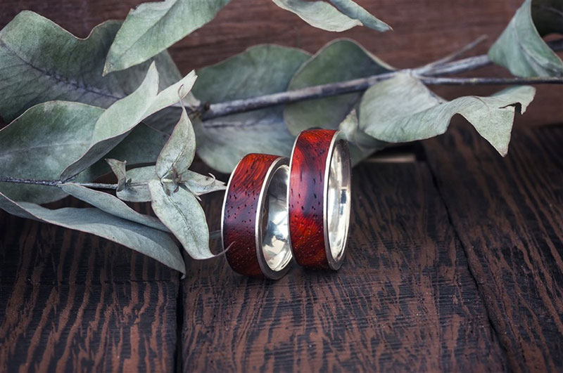 Two men's rings of different sizes, made of wood and silver, resting on a rustic wooden surface beside dried eucalyptus