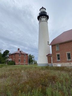 A white lighthouse surrounded by two red-brick buildings, set against a cloudy sky