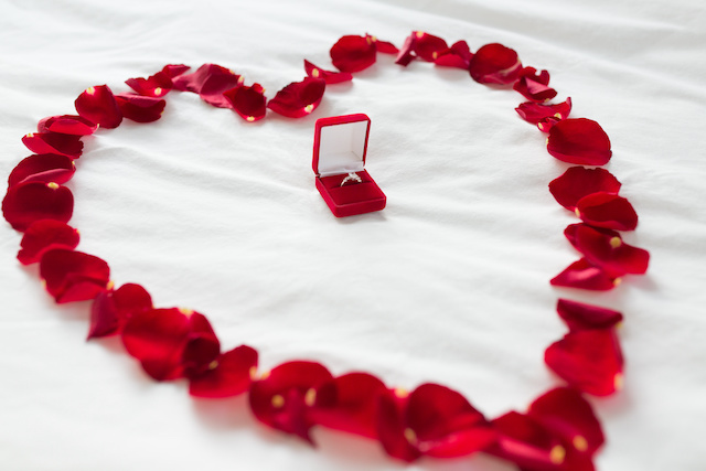 Red ring box with diamond ring inside, surrounded by heart petals in the shape of a heart on bed sheets