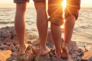 A close-up of a couple's legs standing on a rocky beach, enjoying a picturesque sunset with warm hues