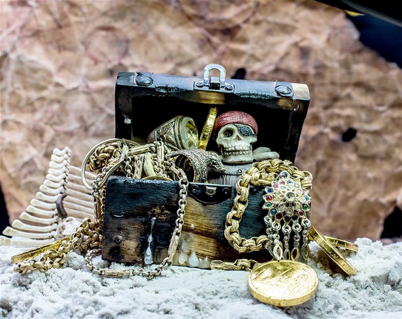An open pirate's treasure chest filled with assorted gold jewelry and coins, placed on a coral-strewn beach