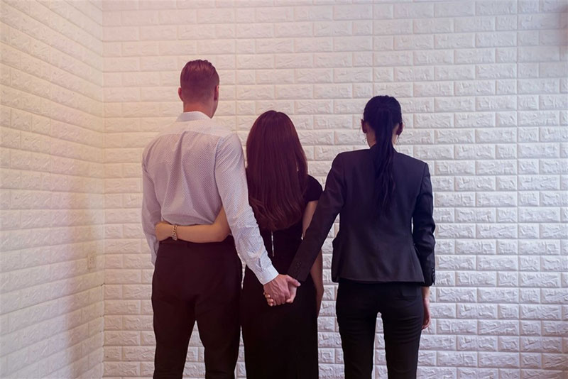 Three people standing together in front of a white wall, suggesting themes of betrayal and unfaithfulness