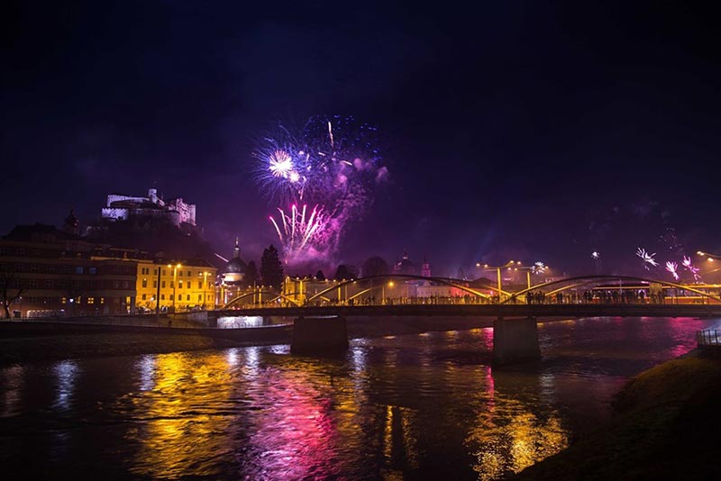Colorful fireworks illuminate the night sky above a river, with a bridge prominently featured in the scene