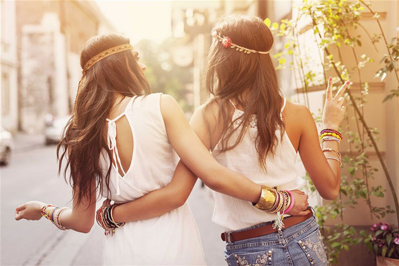 Two women dressed in hippie-inspired clothing and jewelry, with their backs turned, exuding a joyful and warm atmosphere