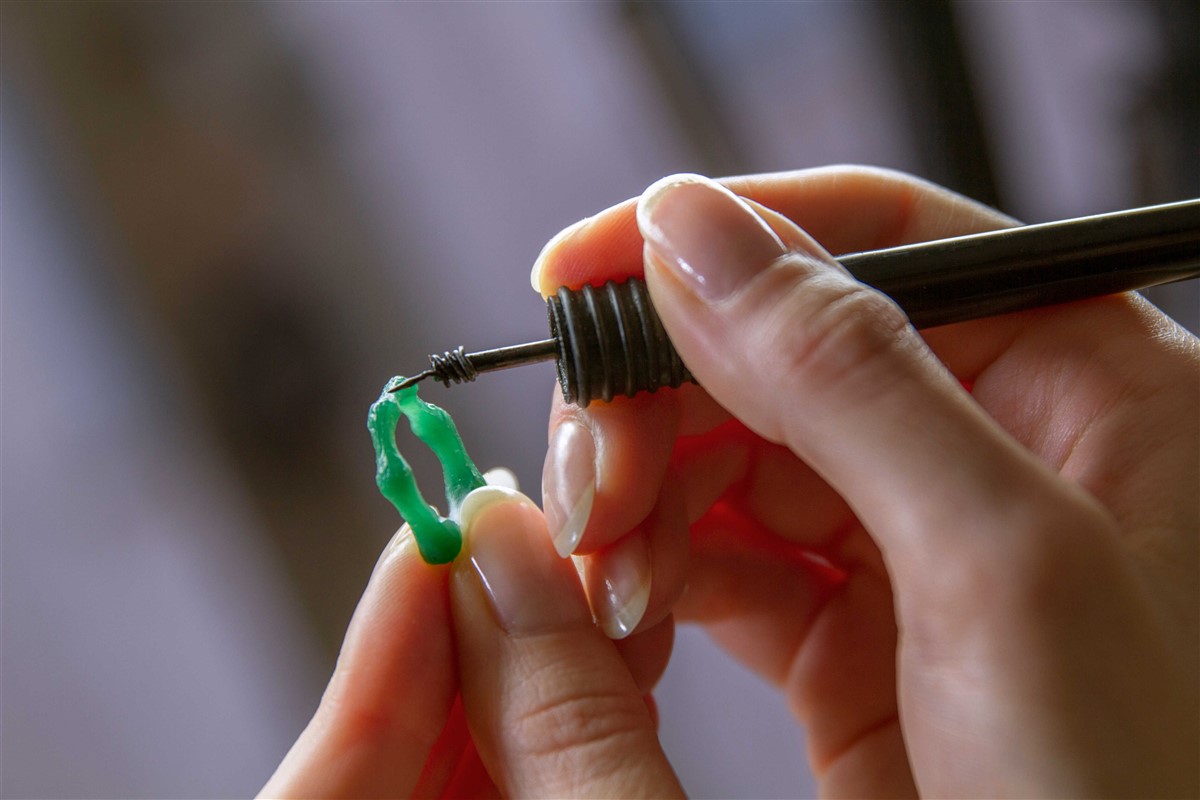 A close-up of hands intricately sculpting a green wax model of a ring with a tool