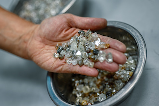 A close-up of rough diamonds on a hand, taken from metal bowls filled with additional rough diamonds visible in the background