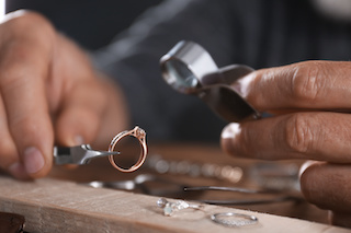 A close-up of a jeweler inspecting a diamond ring with a loupe and tweezers on a wooden table, with various jewelry tools scattered around
