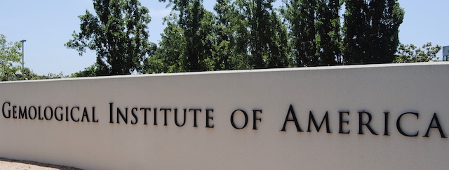 A sign reading "Gemological Institute of America" on a white wall, with tall green trees and a clear sky in the background
