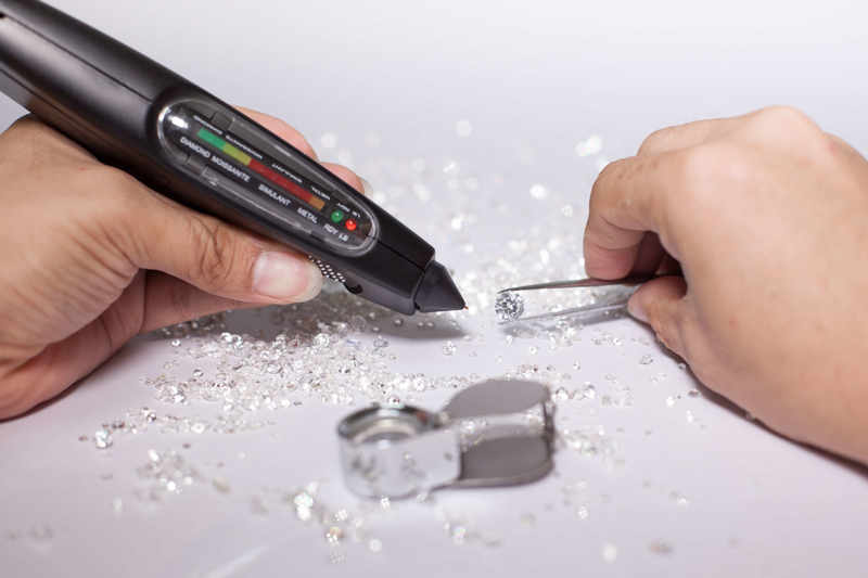 An expert using a handheld diamond tester to examine a diamond held by tweezers on a white surface, with a jeweler's loupe and scattered stones also visible
